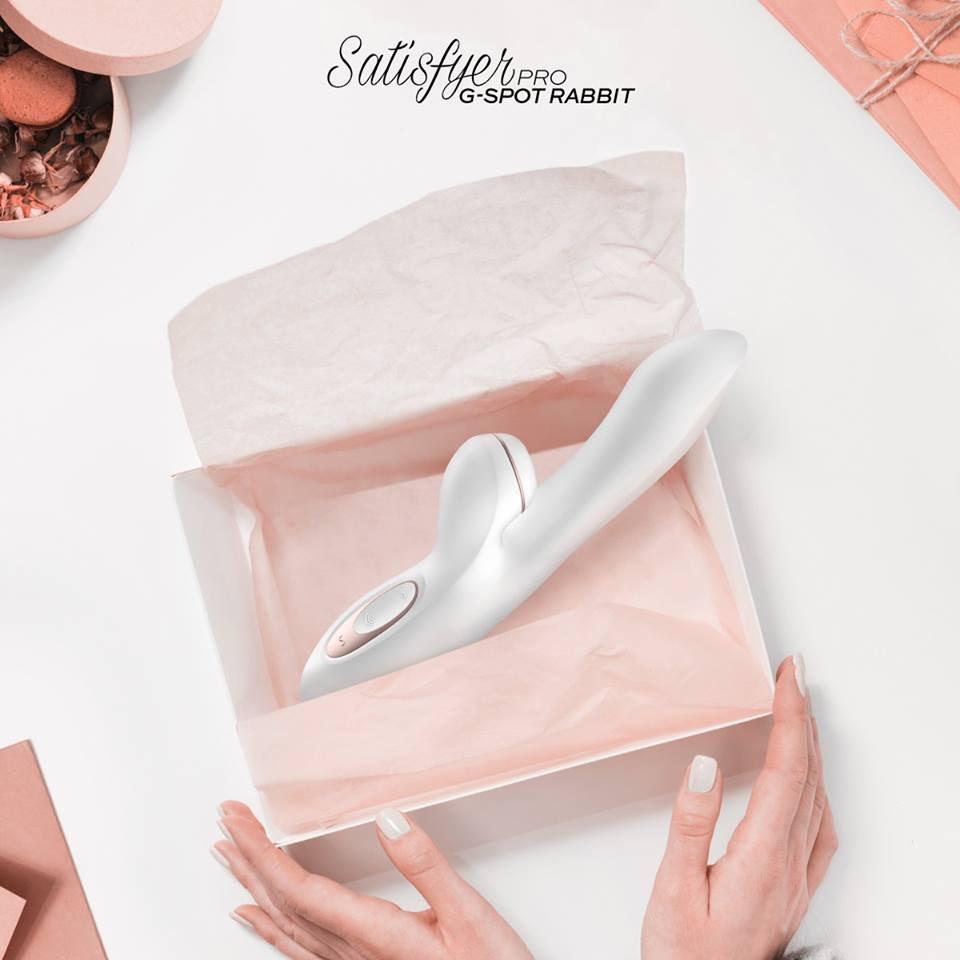 A pink cardboard box filled with pink tissue paper is pictured open with a white long slender banana shaped product within the box and being held by a pair of women's hands.