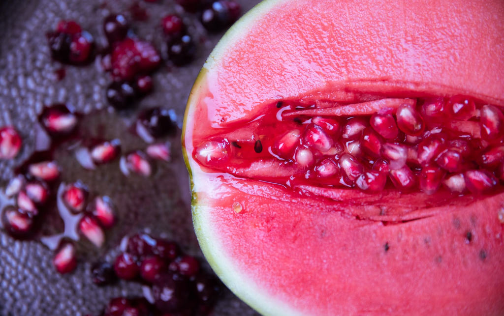 A close up image of a piece of fruit cut in half with it's pink flesh exposed and pink seeds fill up the middle of the slice while some pink seeds are also pictured on the counter beside it.